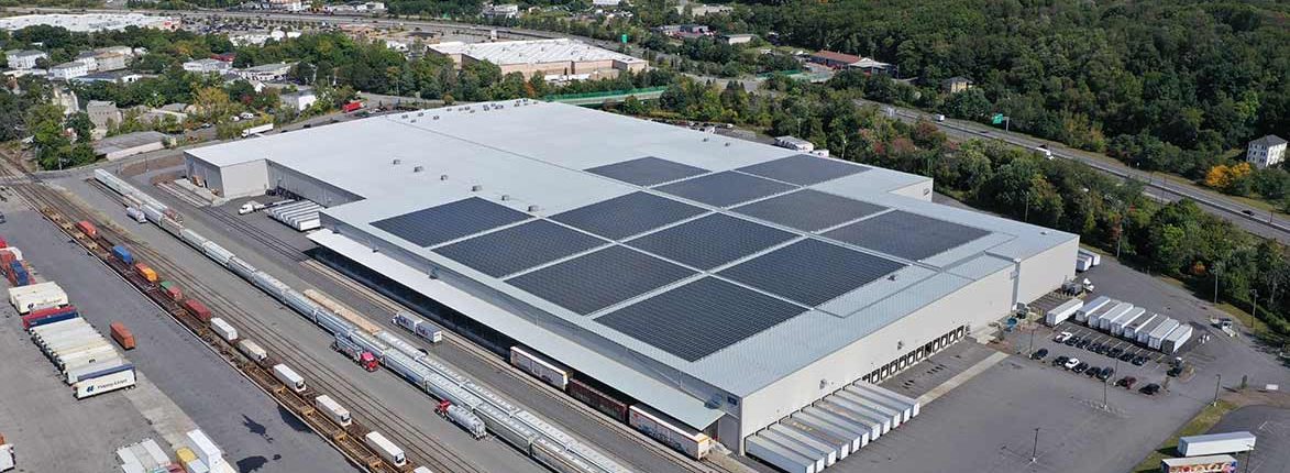 Aerial view of a large industrial warehouse with solar panels on its roof. The surrounding area includes train tracks with several freight trains, parking lots, and dense green trees in the background.