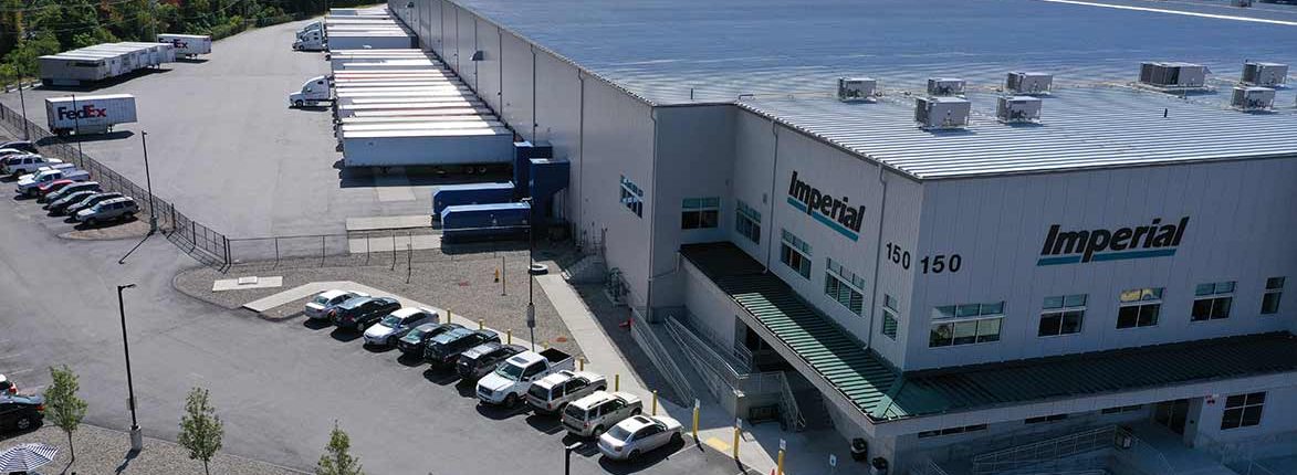 Aerial view of a large industrial warehouse with Imperial signage. Several delivery trucks are parked at loading bays, and cars are in the adjacent parking lot. The facility is surrounded by a fence and trees.