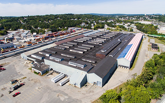 Aerial view of a large industrial warehouse with a roof painted like the American flag. Surrounding the building are trees and a parking area with several vehicles. The setting appears to be a suburban or semi-industrial area.