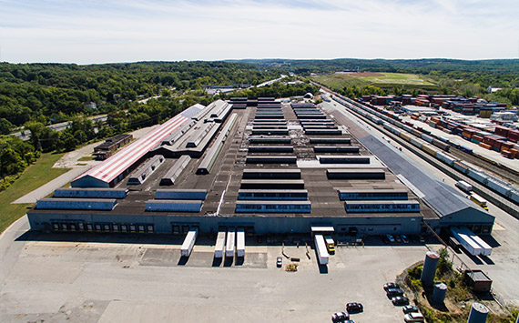 Aerial view of a large industrial warehouse with multiple loading bays and flat roofs. Surrounding the building are trees and a railway with numerous train cars. The sky is clear and the area is expansive.