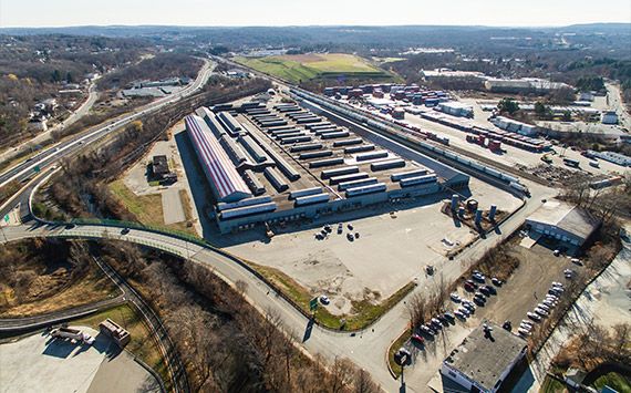Aerial view of a large, rectangular industrial facility with rows of parked buses. Surrounding the facility are roads, greenery, and additional structures. Vehicles are visible in parking lots around the area.