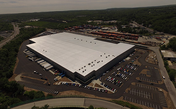 A large warehouse with a white roof surrounded by a parking lot filled with cars. There are multiple trucks docked at the loading bays. The area is surrounded by trees and open land, with a railway line and some containers nearby.