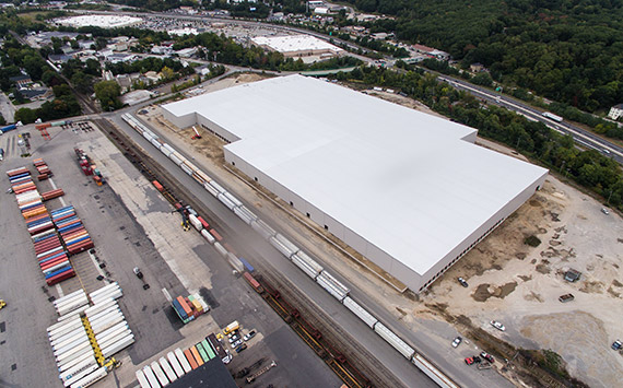Aerial view of a large white warehouse surrounded by trees. Train tracks with several cargo trains run alongside the building. Nearby, there are colorful shipping containers and an industrial area.