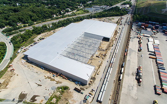 Aerial view of a large warehouse under construction, surrounded by greenery. Rail tracks with stationary trains and shipping containers are visible on the right. A roadway with vehicles runs in the background.