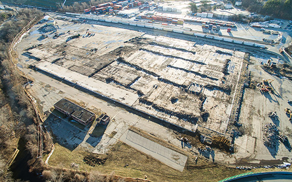 Aerial view of a large construction site with cleared land and exposed foundations. Heavy machinery is visible, and the area is surrounded by trees and a parking lot with numerous vehicles in the background.