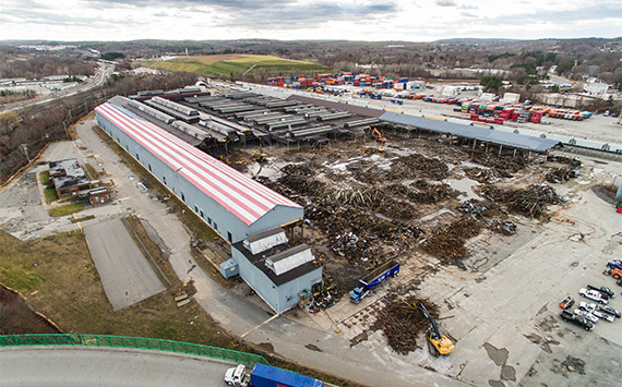Aerial view of a large industrial area with a long building on the left and piles of scrap metal scattered around. Numerous shipping containers are visible in the background, along with roads and a wooded area under a cloudy sky.