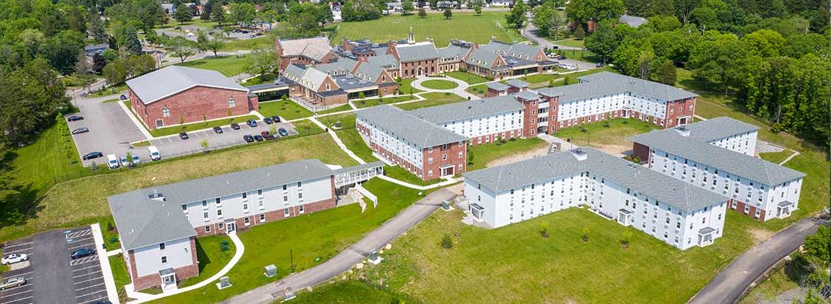 Aerial view of a college campus with several multi-story brick and white buildings arranged in a U-shape. Surrounding the buildings are parking lots and grassy areas with trees. A large building sits in the background near open fields.