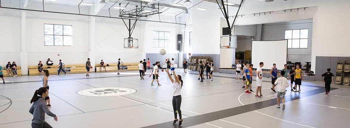 A spacious indoor basketball gym with people engaged in various activities. Some are shooting hoops while others are seated on bleachers. The high ceiling has lights and a basketball hoop. The floor has court markings and a circular logo in the center.