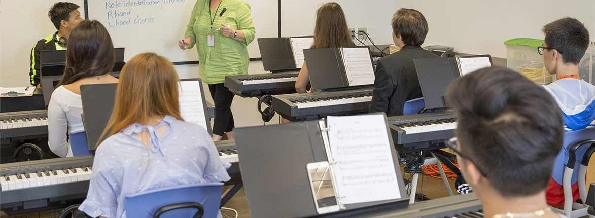 A classroom with students sitting at electronic keyboards facing a teacher, who is standing and speaking near a whiteboard. The students are attentive, with some taking notes. The room is well-lit and organized.
