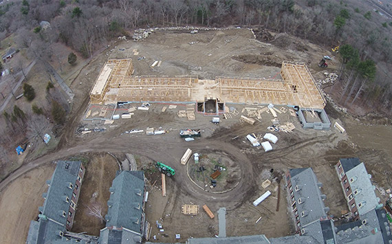 Aerial view of a construction site surrounded by trees. Foundations of a large building are visible, with scattered construction materials and vehicles. Existing buildings are on the lower side of the image. Earth and cleared land dominate the scene.