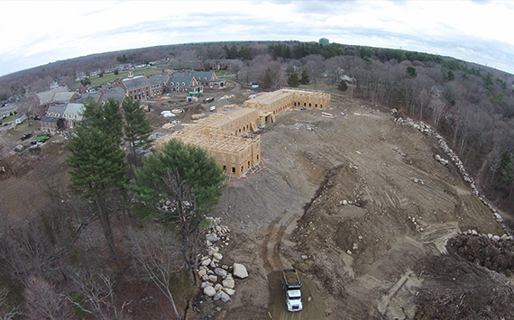 Aerial view of a construction site with a partially built wooden structure surrounded by cleared land and piles of dirt. Nearby, there are residential buildings and dense trees, with a truck parked on the dirt in the foreground.
