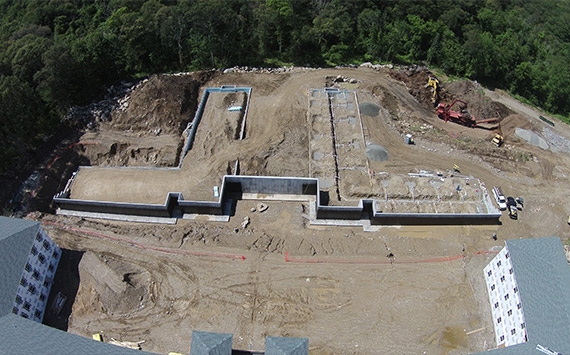 Aerial view of a construction site surrounded by trees. The site shows the foundation of a building in progress, with concrete walls and cleared earth. There are construction vehicles and materials around the perimeter.