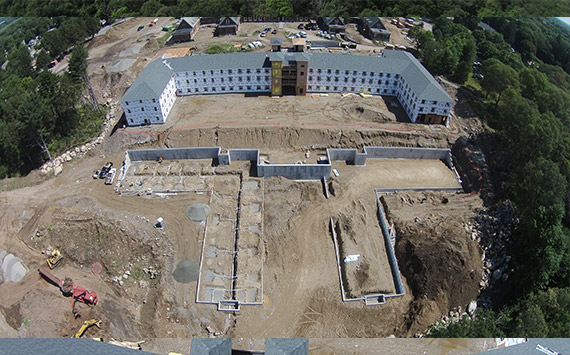 Aerial view of a large construction site surrounded by trees. There is a partially constructed building in a U-shape, with foundations and walls in progress. Construction vehicles and materials are scattered around the site.