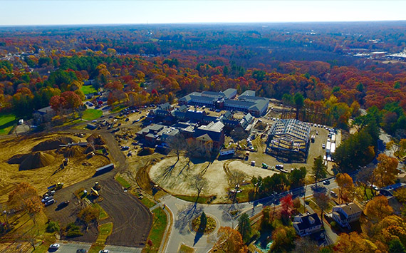 Aerial view of a construction site surrounded by autumn trees. There are multiple buildings, construction vehicles, and a steel framework structure. Roads and houses are visible in the foreground, and a dense forest is in the background.