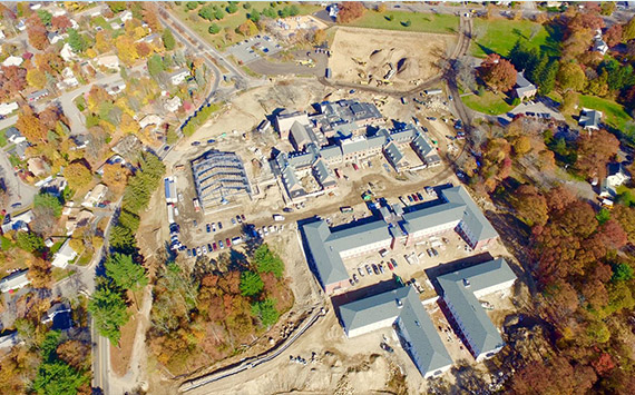 Aerial view of a construction site surrounded by autumn trees. Several large buildings are under development with construction vehicles scattered around. Residential houses and roads are visible in the upper left corner.
