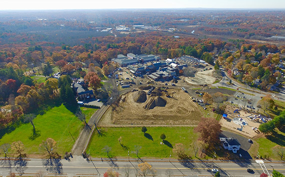 Aerial view of a construction site with piles of dirt surrounded by trees with autumn foliage. Roads and buildings are visible nearby, with a clear sky in the background.