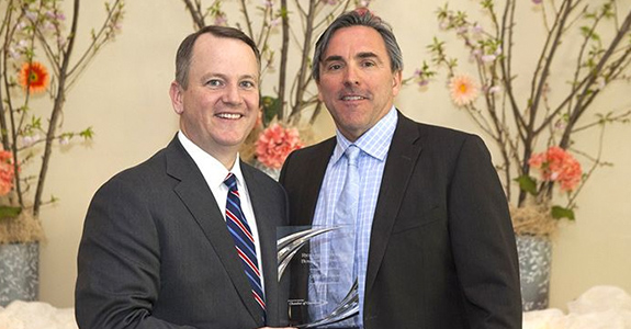 Two men in suits stand side by side, smiling. One man holds an award with a curved design. In the background, there are vases with branches and flowers, creating a decorative setting.