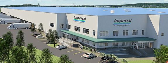 A large industrial warehouse with a white exterior and the name Imperial displayed on the building. Several trucks are parked by loading docks, and a few cars are in the parking lot. Trees and grassy areas surround the facility.