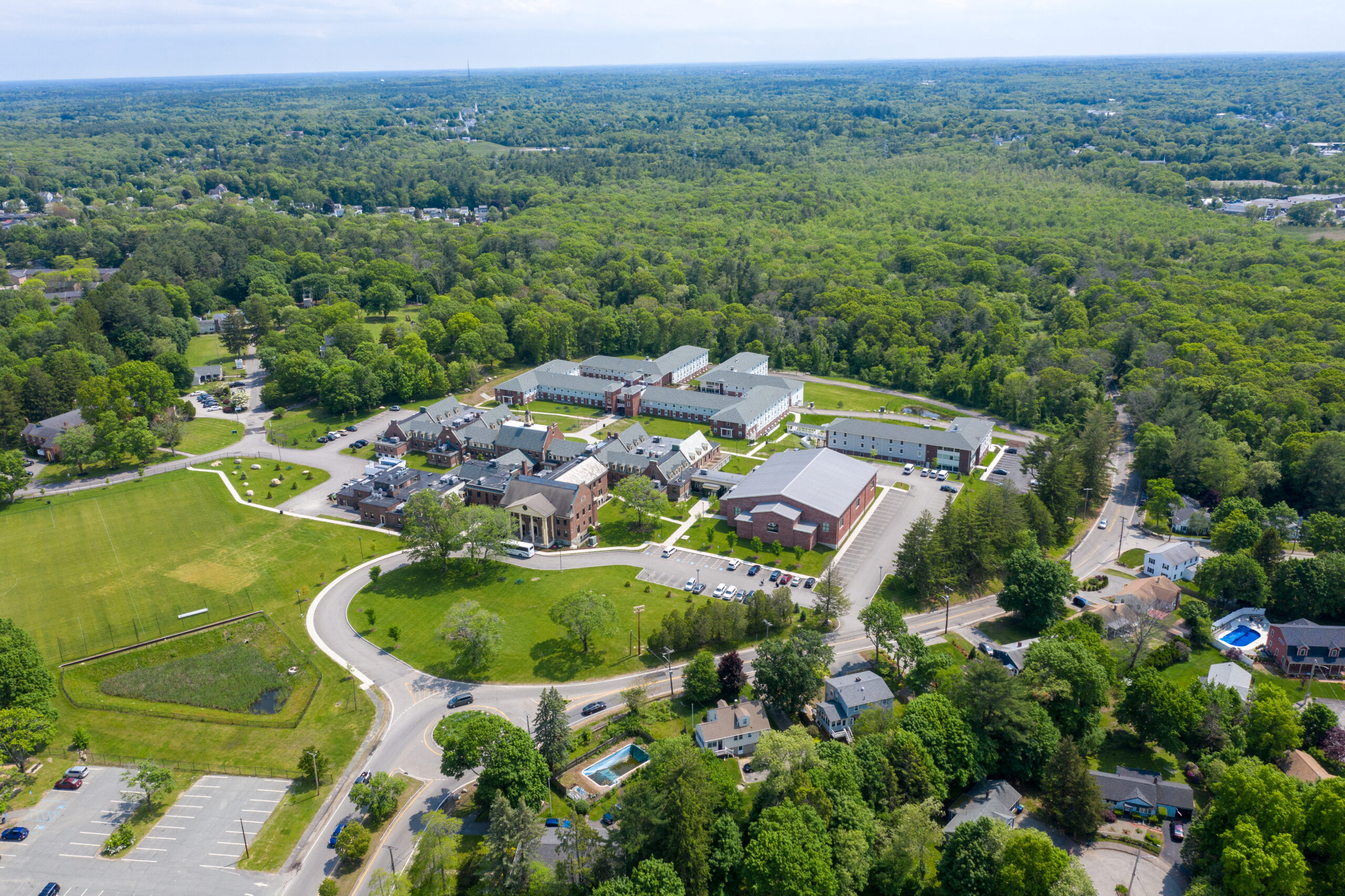 Aerial view of a campus with multiple buildings surrounded by greenery and trees. Roads and pathways connect the structures. Residential houses and a sports field are visible nearby, with a dense forest in the background.