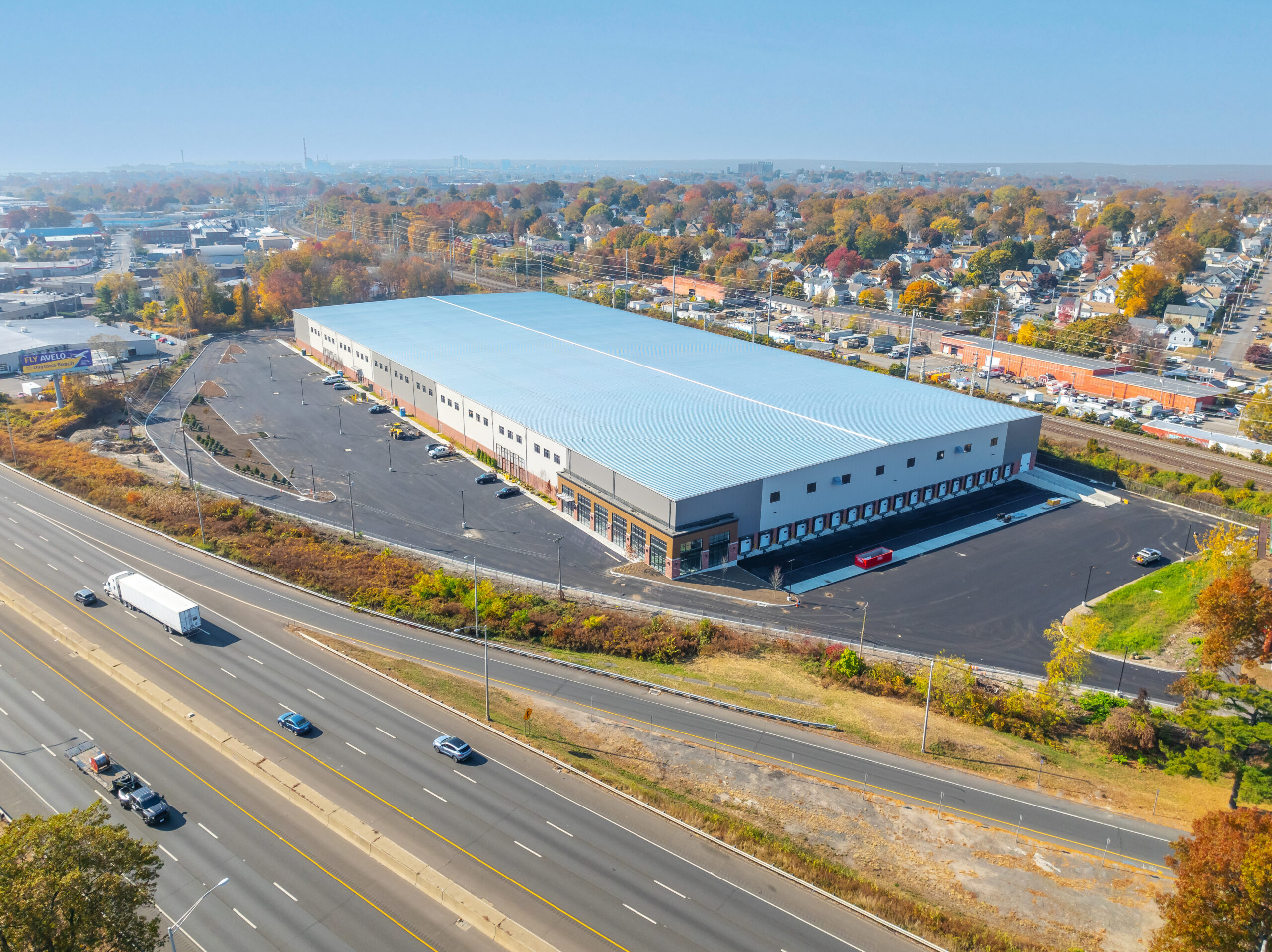 Aerial view of a large warehouse with a light blue roof, surrounded by a spacious parking area. The building is located near a highway with several vehicles. In the background, theres a suburban area with houses and trees.