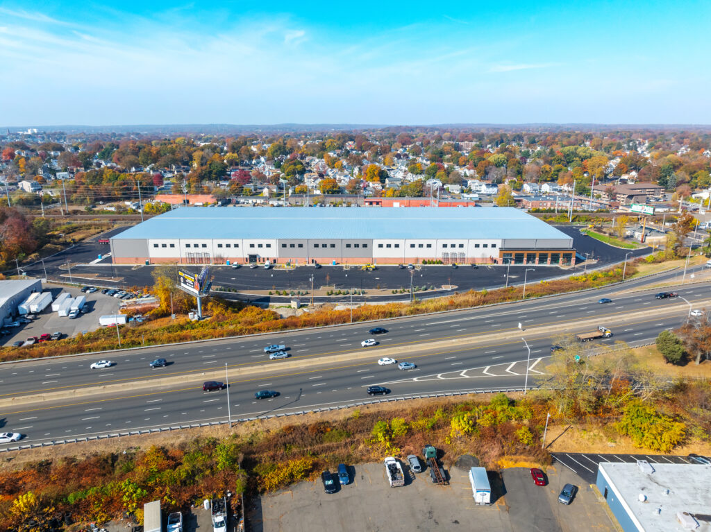 Aerial view of a large industrial warehouse with a gray roof and multiple loading bays, situated next to a busy highway with several cars. In the background, a suburban area with autumn-colored trees is visible under a clear blue sky.