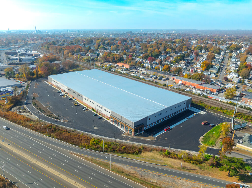 Aerial view of a large rectangular warehouse with a blue roof, surrounded by a parking lot and roads. The area features autumn foliage, residential houses, and a highway in the foreground. The sky is clear and blue.