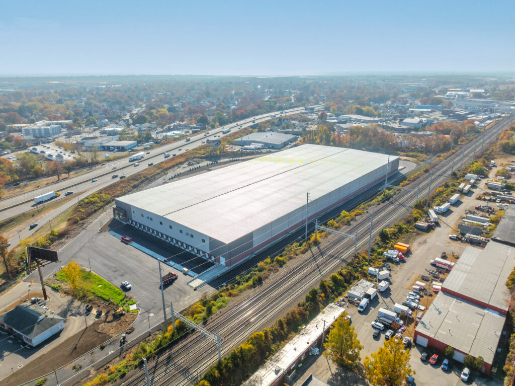 Aerial view of a large industrial warehouse with a flat roof surrounded by parking lots and train tracks. Nearby roads and highways are visible, with a sprawling cityscape in the background under a clear sky.