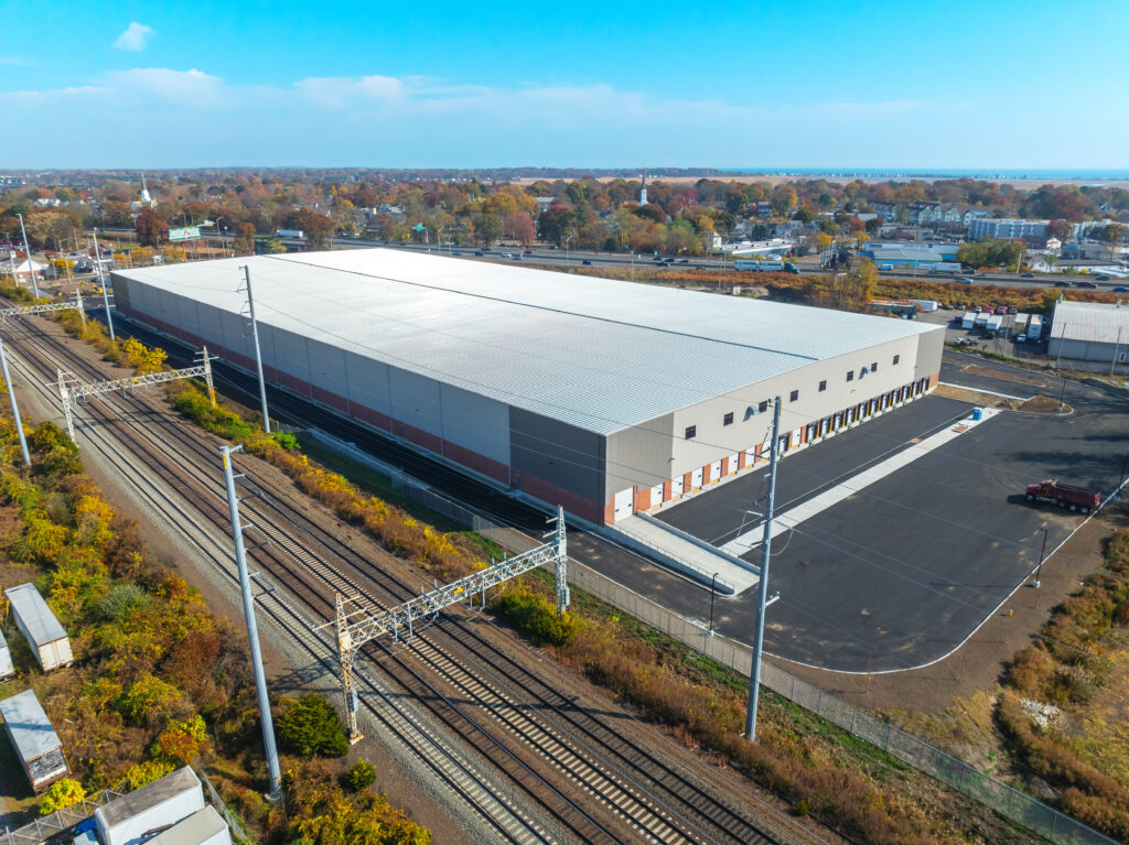 Aerial view of a large warehouse with a white roof and gray walls. Nearby are railway tracks and surrounding greenery. In the distance, there are residential and industrial buildings under a blue sky.