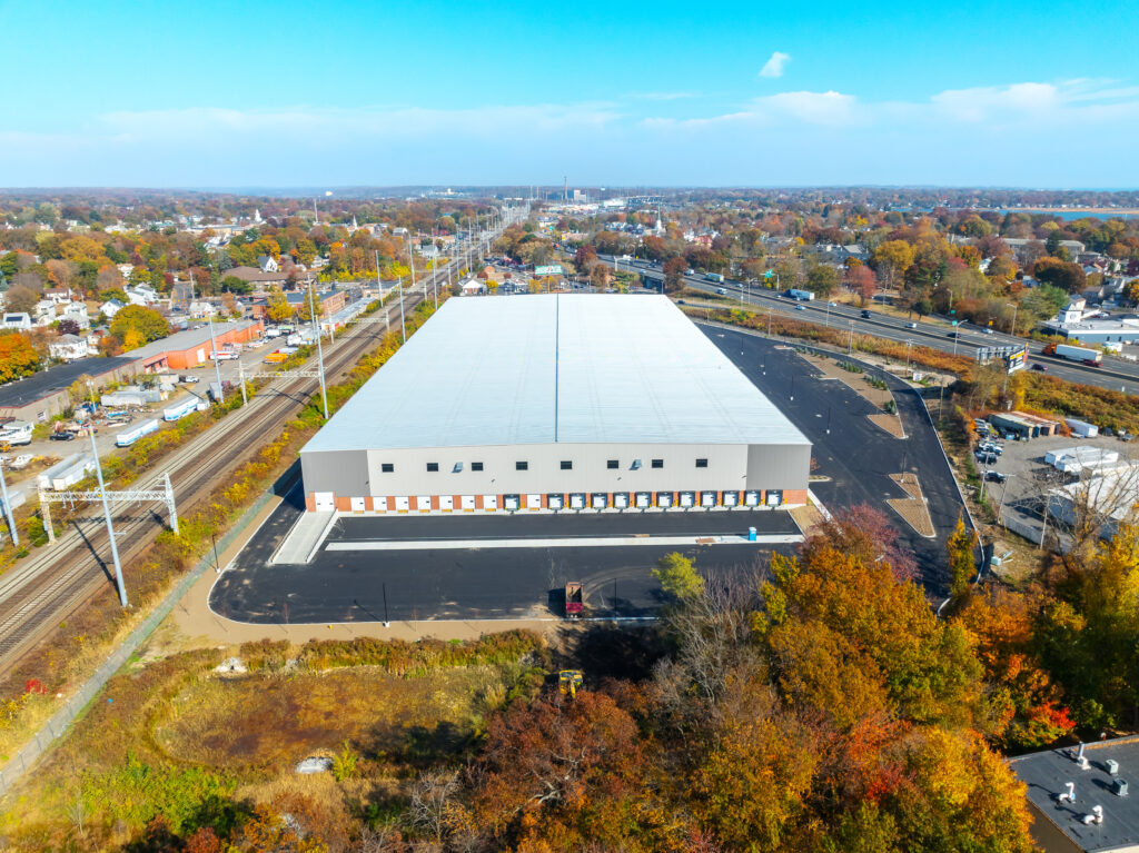 A large white warehouse with multiple loading docks, surrounded by autumn trees and adjacent to railway tracks. The view includes nearby roads, scattered buildings, and a clear blue sky overhead.