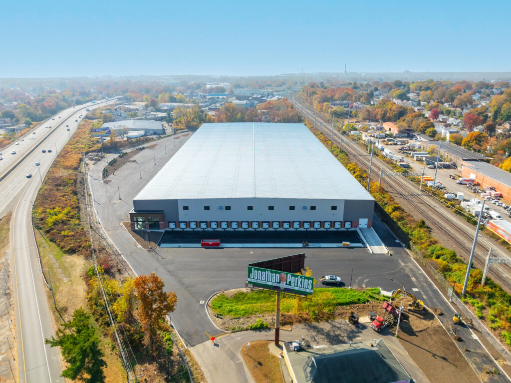 Aerial view of a large, rectangular warehouse with a light gray roof, surrounded by parking and entry areas. Its situated near a highway and train tracks, with an advertising billboard visible in the foreground. Trees with autumn foliage line the scene.