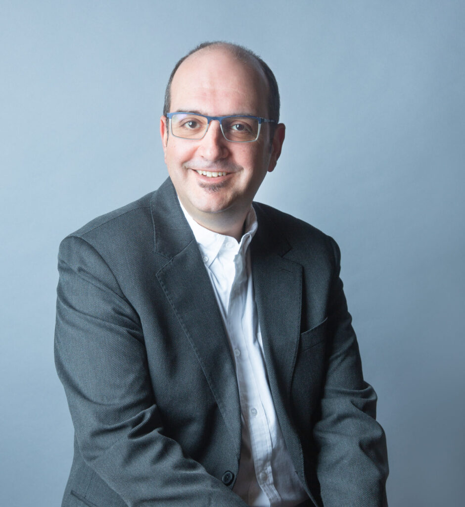 A man wearing glasses and a dark suit with a white shirt sits on a stool, smiling at the camera against a plain light blue background.